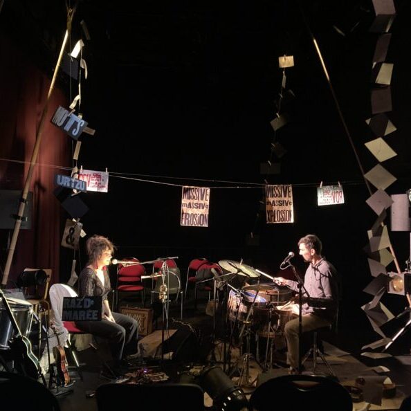 Coline et Clément se font fasses, assis. Il et elle sont de profil et leurs instruments les séparent ; une guitare électrique, une shruti, des clochettes, une batterie. Un fil traverse l'image au dessus d'eux, sur lequel sont accroché des papiers typographiés. De grands bambous suspendent des guirlandes de feuilles blanches et colorées dessinant des lignes horizontales et diagonales.